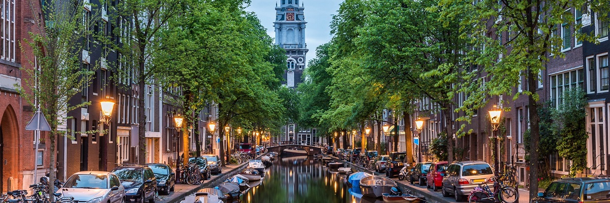En kanal i Amsterdam kantad av b�tar, bilar cyklar och bostadshus. L�ngst bort ligger sticker ett kyrktorn upp som speglar sig i vattnet.