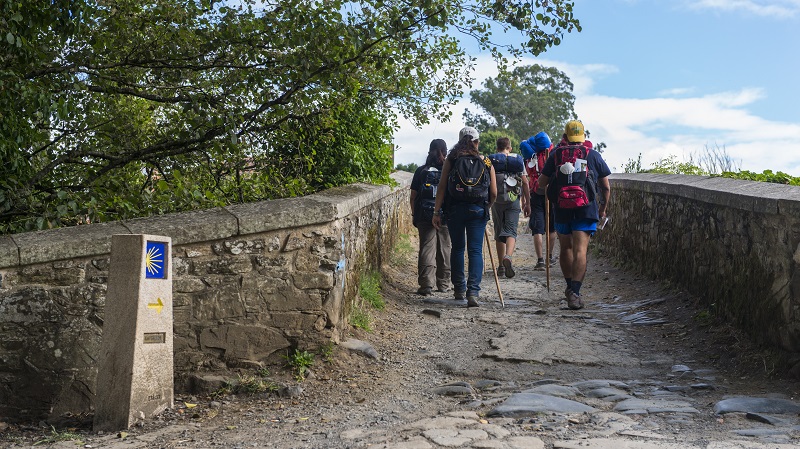 En grupp pilgrimsvandrare gr bortt ver en bro, ledsagade av den gula pilgrimsmusslan p en lg stenpelare.